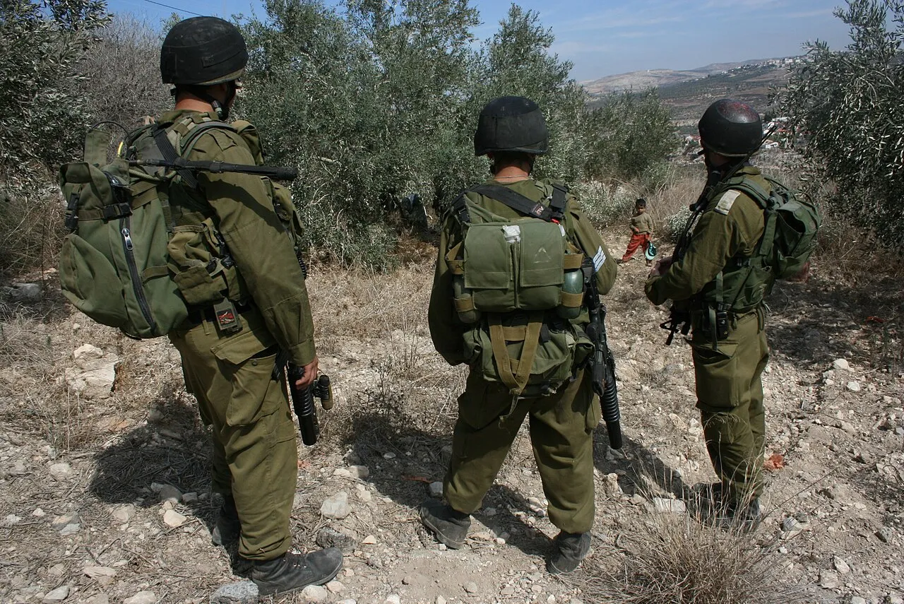Soldaten staan tussen de olijfbomen op de Westelijke Jordaanoever tijdens de olijfoogst, met een Palestijnse jongen voor hen